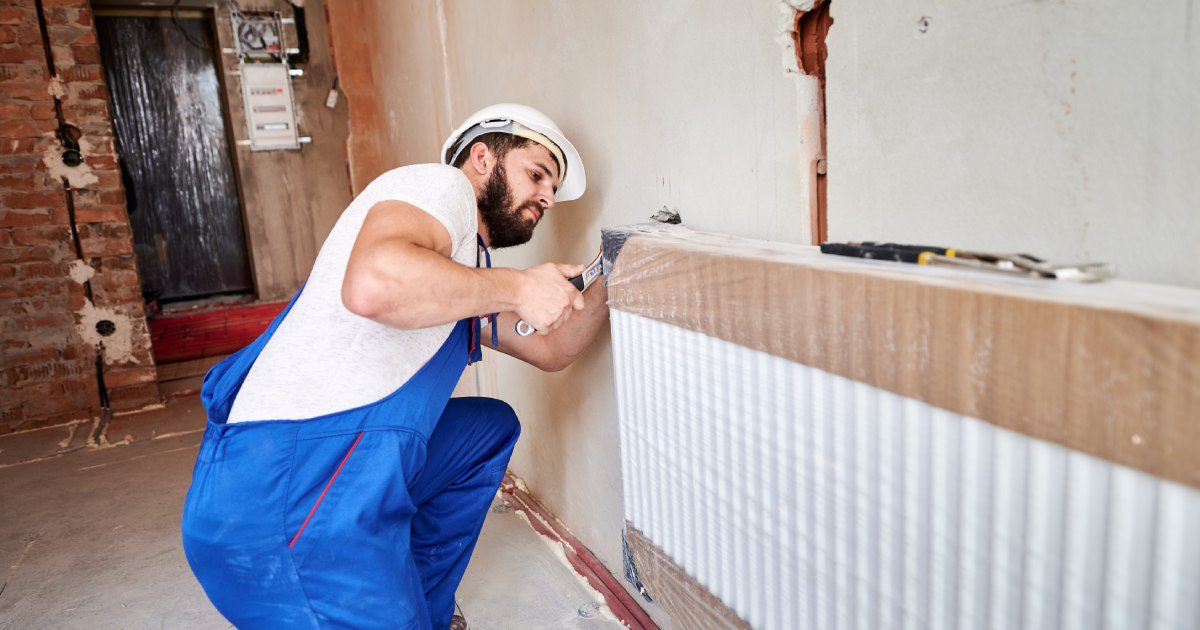 Young handsome plumber installing water radiator in unfinished apartment | Do Plumbers Install Central Heating